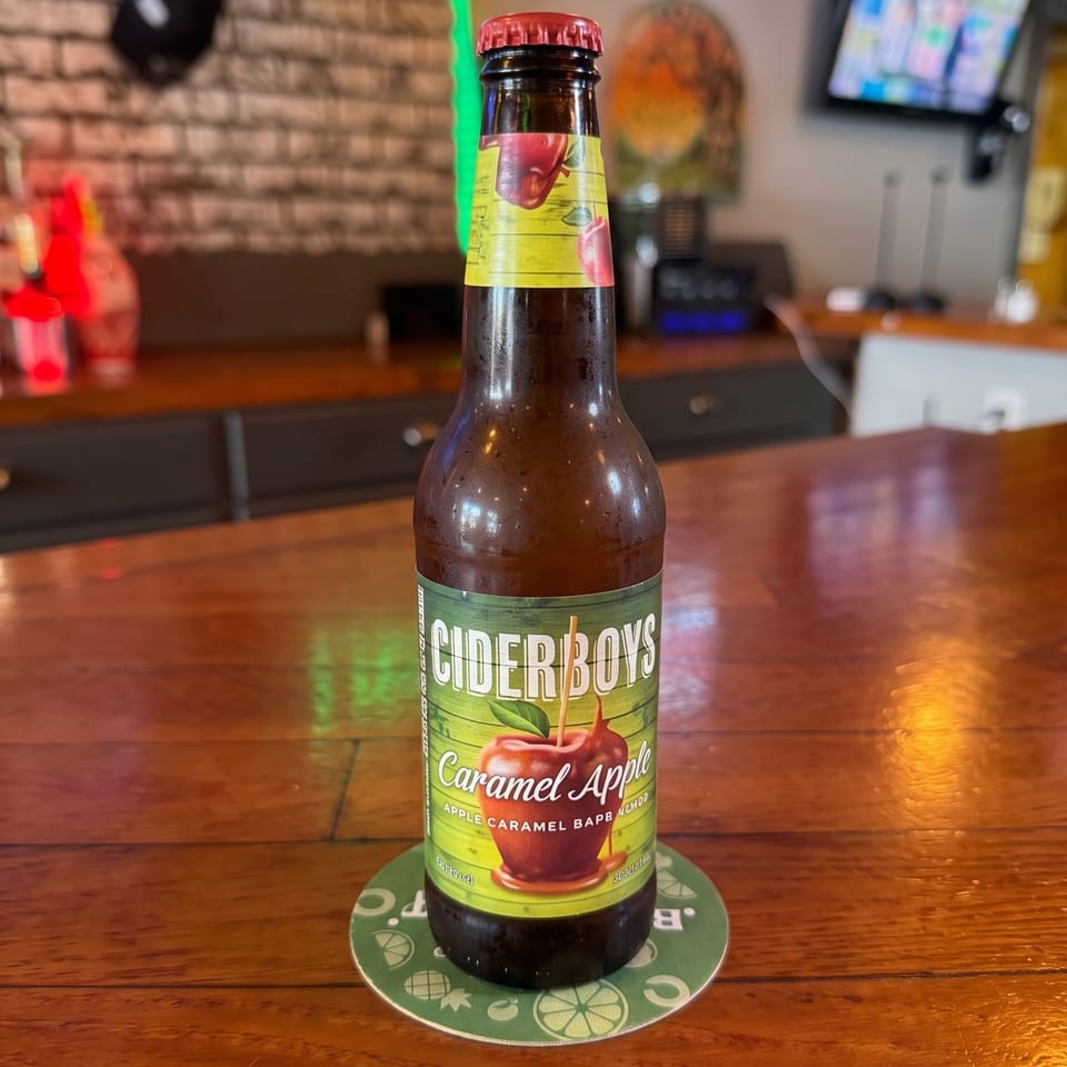 Ciderboys Caramel Apple cider bottle on a coaster at a warmly lit bar, with neon signs, televisions, and brick walls in the background.