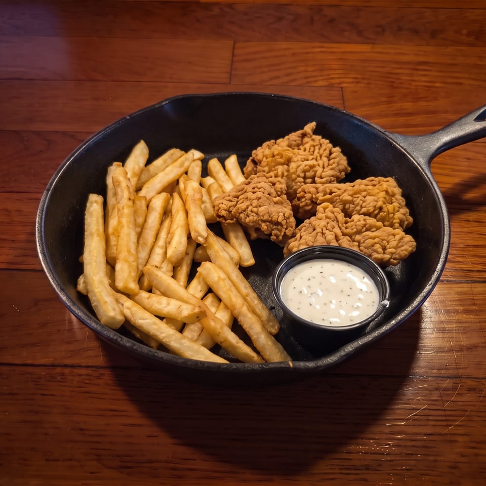 Cast-iron skillet filled with crispy chicken tenders, French fries, and a cup of creamy dipping sauce on a wooden table.