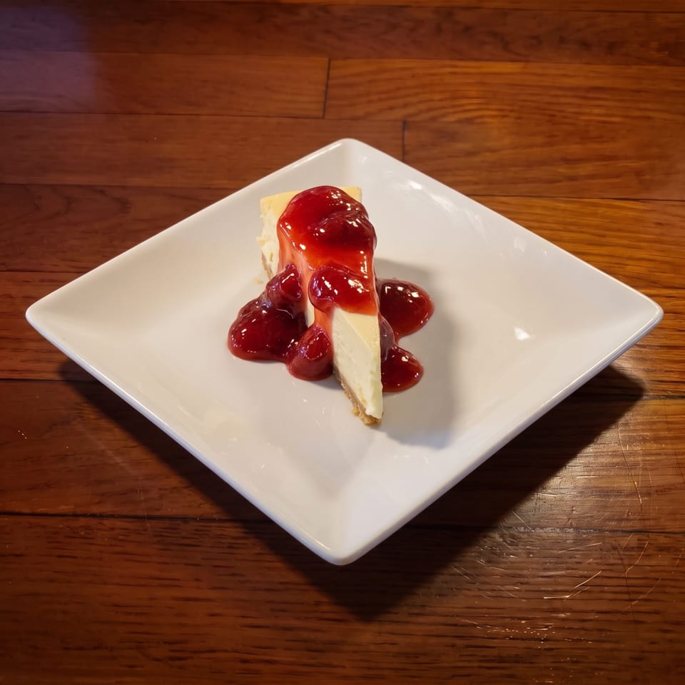Slice of creamy cheesecake topped with glossy red berry sauce on a white square plate.