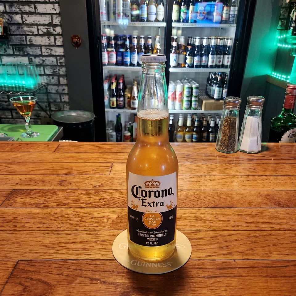 Corona Extra beer bottle on a wooden bar, with a refrigerated display of assorted beverages, cocktail, and condiments behind it.
