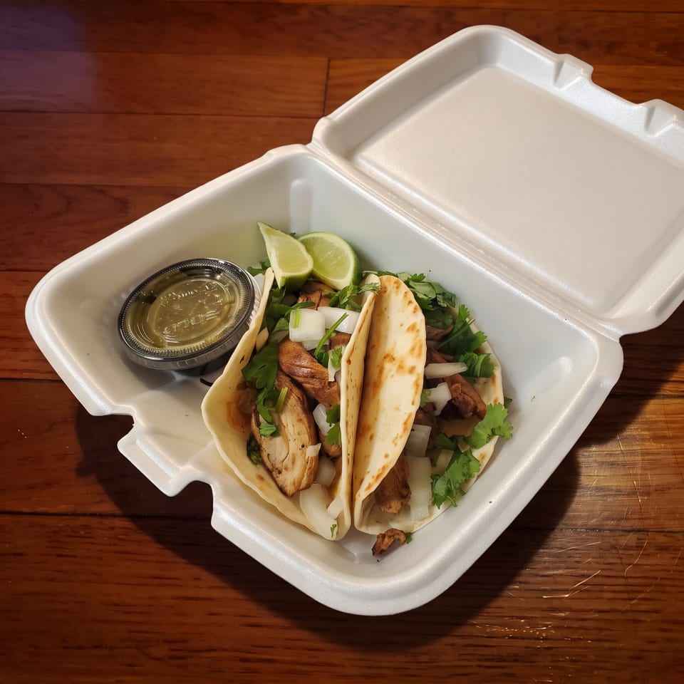 Two tacos filled with grilled meat, onions, and cilantro in a takeout container with lime wedges and dipping sauce.