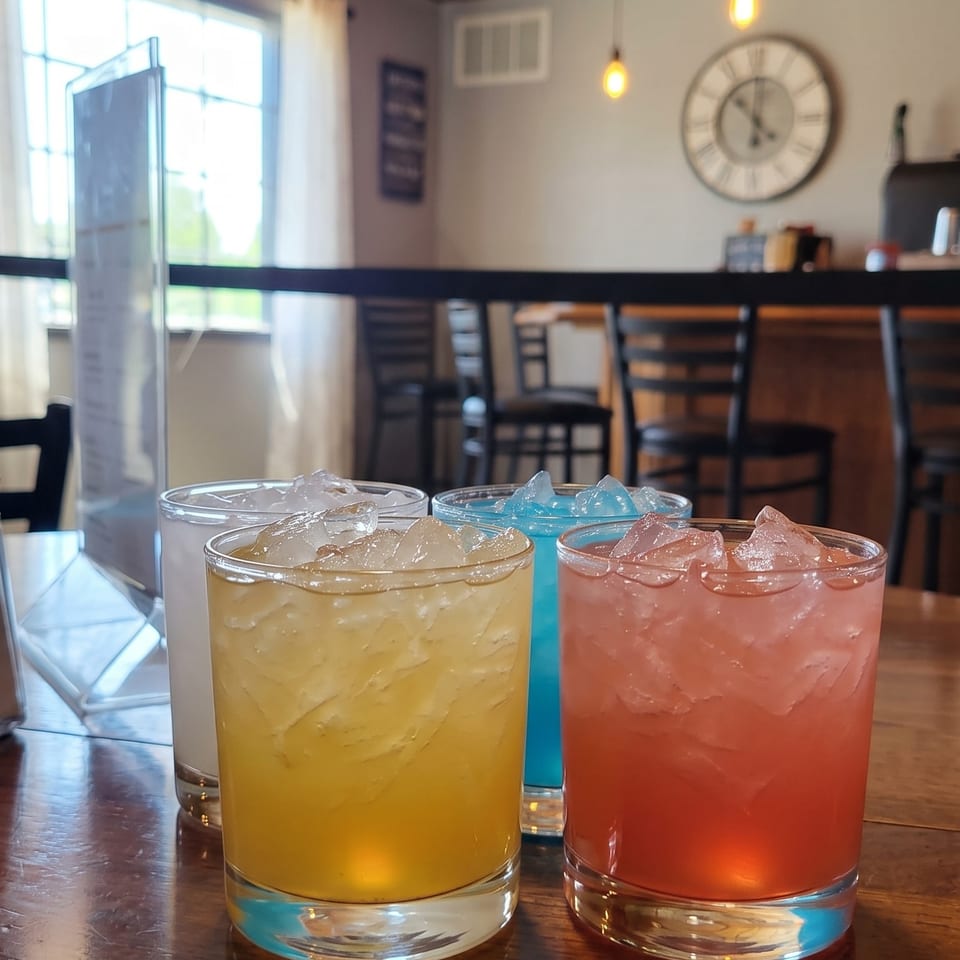 Four colorful iced drinks sit on a wooden restaurant table beneath pendant lights, with a large wall clock and chairs in the background.