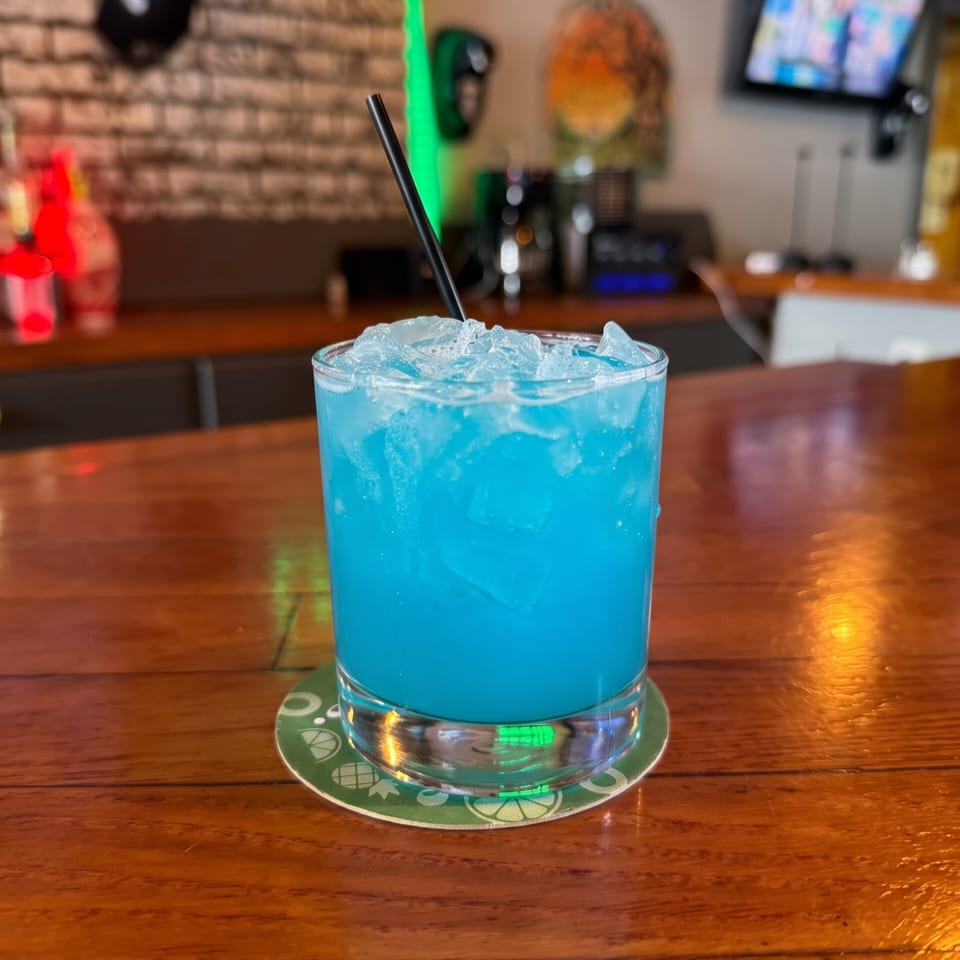 Bright blue iced cocktail with a black straw on a branded coaster at a warmly lit bar.