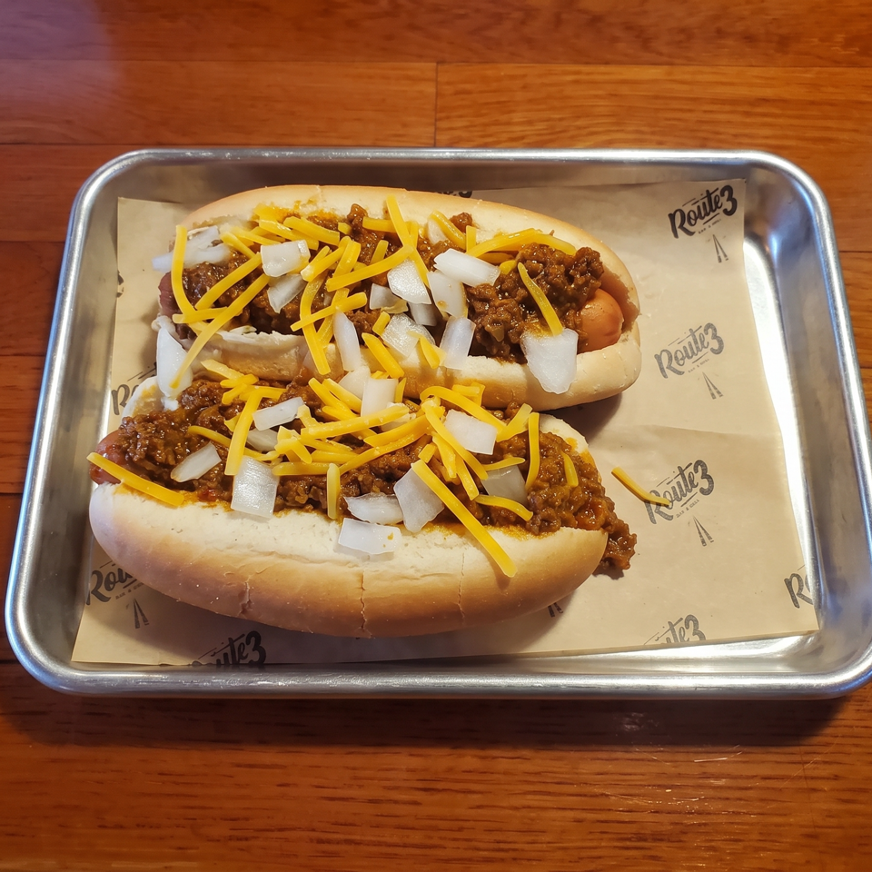 Two chili dogs topped with shredded cheddar cheese and diced onions, served in a metal tray on a wooden table.