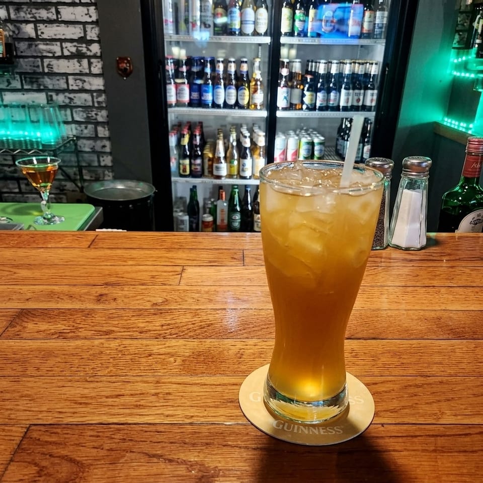 A chilled amber drink with ice and straw sits on a Guinness coaster at a wooden bar, with bottled beers displayed behind it.