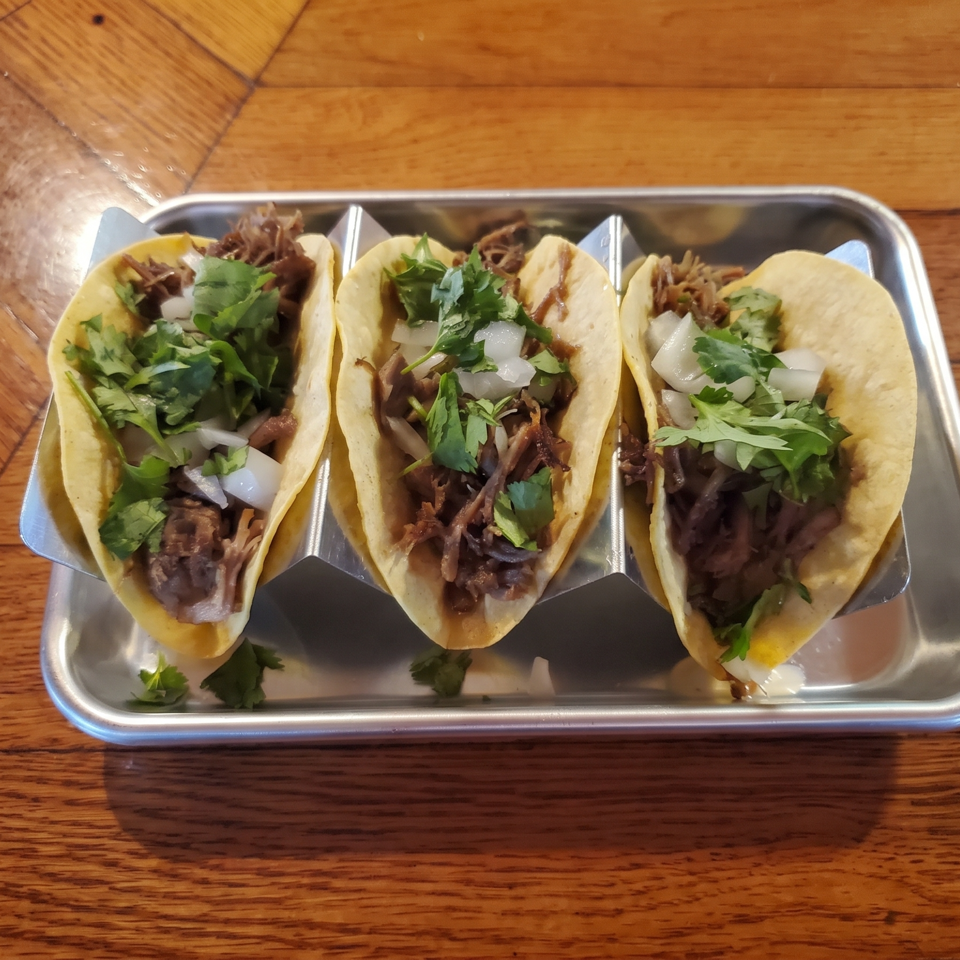 Three soft tacos filled with shredded meat, diced onions, and cilantro on a metal tray atop a wooden table.